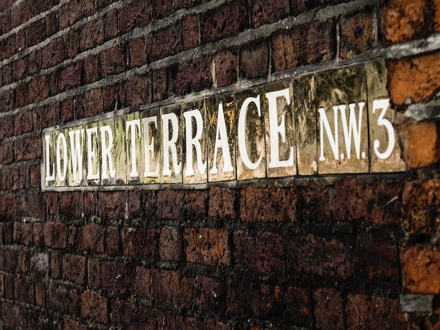 Close-up view of a vintage street sign mounted on a weathered red brick wall, displaying the text 'Lower Terrace NW3' in white lettering on a tarnished metallic background. The bricks have a rough texture and are slightly uneven, with some mortar visible between them. The sign appears aged, with signs of rust and moss growth, suggesting exposure to outdoor elements. Soft natural lighting highlights the textured surface of the bricks and the reflective metal of the sign, providing a clear visual of the urban environment associated with the West End Lane area in West Hampstead. This setting emphasizes the importance of regular surface cleaning and maintenance, often provided by [COMPANY_NAME], for preserving historic signage and urban aesthetics in residential or commercial spaces.