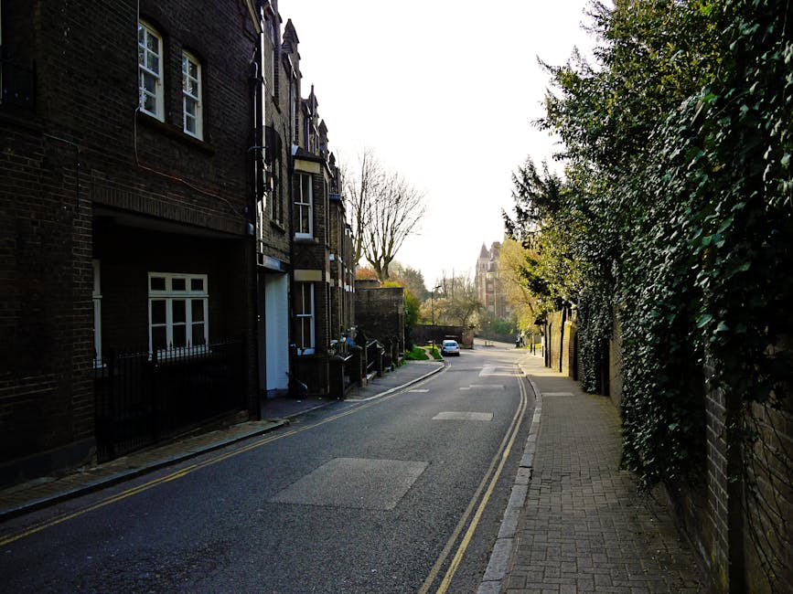 A narrow residential street in West Hampstead with brick buildings on the left side featuring white-framed windows and black metal railings. The street is paved with asphalt, displaying double yellow lines along the curb, and a concrete sidewalk runs alongside the buildings. On the right side, there is a tall, dense hedge and a brick wall partially covered with greenery, with a small section of the pavement visible. In the background, trees with sparse foliage and a distant church steeple are visible under a bright, partly cloudy sky, suggesting early spring or late autumn. The scene is well-lit with natural daylight, emphasizing the cleanliness of the area, and the overall atmosphere is quiet and tidy, suitable for domestic and commercial surface cleaning discussions related to basement flat access issues on West End Lane.