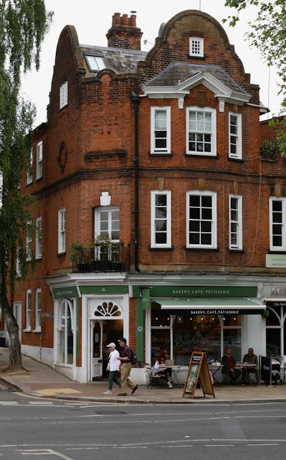 An old brick wall with moss and weathering, featuring a metal grate arch at its base and a street sign indicating directions to Branch Hill and West Heath Road. Behind the wall, a large tree with some visible branches and a textured bark trunk stands, with surrounding residential buildings made of brick and tiled roofing. The scene is outdoors with natural lighting, displaying a partly overcast sky. Cleanliness is not applicable as the image focuses on the outdoor structure and natural elements; however, the wall and nearby area appear to be free of litter or debris. This setting exemplifies the historic urban landscape near West Hampstead station, and is mentioned in the context of surface cleaning and maintenance by westhampsteadcarpetcleaning.co.uk for a comprehensive approach to property upkeep.