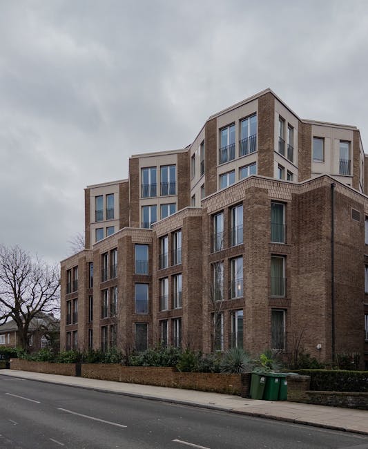 An exterior view of a multi-story residential building in Fortune Green, West Hampstead, featuring a combination of brick and light-colored materials. The building has large, rectangular windows with some sliding doors leading to balconies and terraces, all set against a cloudy sky. The ground level shows a landscaped area with shrubs and small trees, bordered by a low brick wall. A paved sidewalk runs along the street in front, which has two green waste bins positioned at the edge. The overall appearance is clean and well-maintained, with no visible dirt or debris, emphasizing the upkeep of the property. As part of the projects by westhampsteadcarpetcleaning.co.uk, this image illustrates a residential setting where professional cleaning services in the area support the upkeep of both interior and exterior spaces.