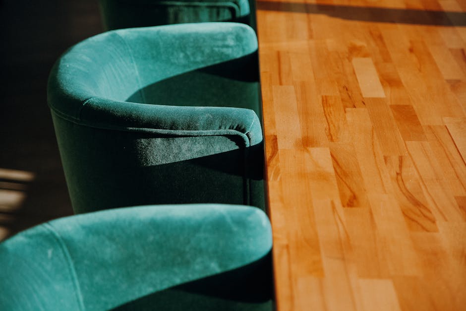 Close-up view of two green upholstered dining chairs with curved backs and plush fabric, positioned alongside a polished wooden dining table with a smooth, shiny surface. The table's surface displays a rich, warm hue with visible wood grain patterns. Natural light illuminates the scene, highlighting the clean and well-maintained condition of both the fabric and the wood, with no visible dust or stains. The setting appears to be a domestic dining area, reflecting a tidy and hygienic environment as part of domestic cleaning services offered by westhampsteadcarpetcleaning.co.uk, focusing on surface cleaning and maintenance.