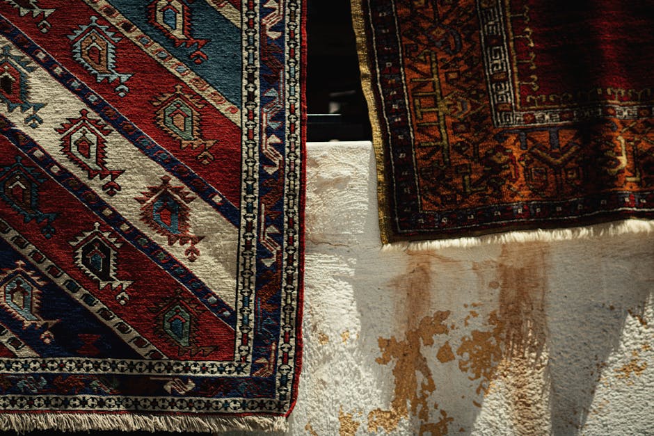 Close-up image showing two traditional area rugs with intricate geometric and floral patterns, laid out on a white textured surface that appears to be a concrete or plaster floor. One rug features a dominant red, blue, and cream colour scheme, while the other displays darker shades of red, orange, and navy. The rugs are partially overlapping, with the edges slightly lifted, revealing the clean, dry surface beneath. Natural sunlight illuminates the scene, highlighting the detailed craftsmanship and vibrant colours of the rugs, suggesting a professional deep cleaning process in a domestic setting. The image emphasizes the importance of surface cleaning and maintenance in preserving the appearance and hygiene of area rugs, with [COMPANY_NAME] providing expert cleaning and care tips for Hampstead Heath area rugs.