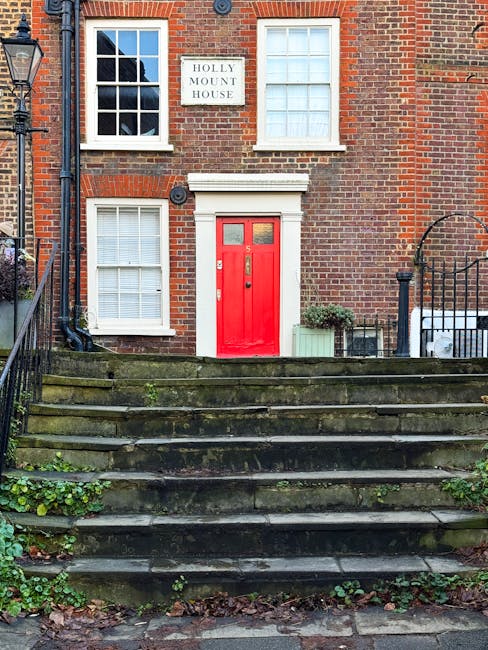 The image depicts the front exterior of a brick residential building with a staircase leading up to a vibrant red front door. The building features four white-framed windows, two above the door and two on the lower level, all fitted with white blinds. A white sign with black lettering reads 'HOLLY MOUNT HOUSE' and is mounted between the upper windows. To the right of the door, there is a small green plant in a pot and a neatly trimmed shrub. The stone staircase appears aged with moss and small plants growing between the steps, indicating outdoor wear. A black metal railing runs along both sides of the staircase. The scene is evenly lit with natural daylight, emphasizing the cleanliness and well-maintained appearance of the exterior, consistent with high-quality surface cleaning and maintenance practices, as exemplified by services such as those offered by westhampsteadcarpetcleaning.co.uk.