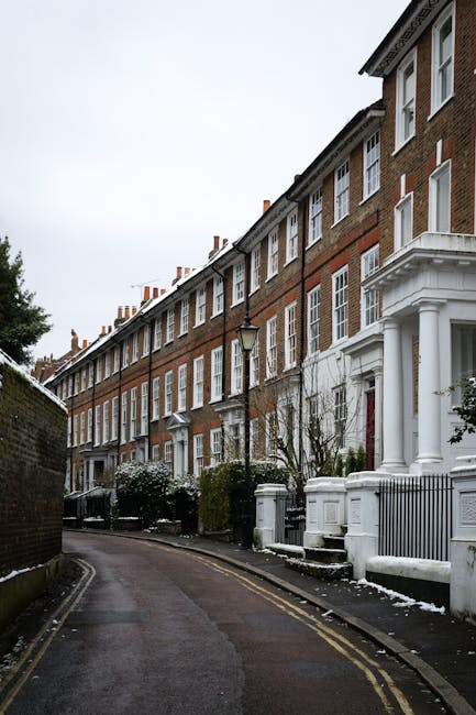 A row of traditional brick residential buildings on a curved street in West Hampstead, NW6, with white-painted doors and decorative columns at the entrances. The street is lined with black iron lampposts and small gardens with bushes and trees, some of which have light snow cover. The ground appears wet, possibly from recent cleaning or weather conditions, and the overall scene emphasizes the clean, well-maintained appearance of the area, reflecting the environment targeted by West Hampstead Carpet Cleaning's surface cleaning and domestic hygiene services.