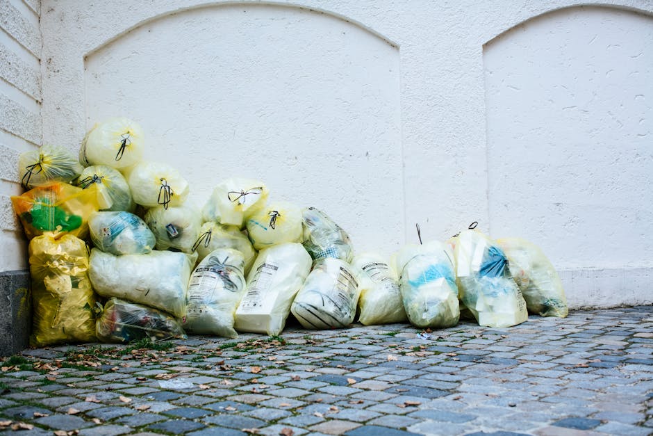 A group of yellow and transparent plastic waste bags filled with trash, stacked against a white textured exterior wall on a cobblestone street. The bags contain various household waste and are tied with black plastic ties, placed near a brick corner with visible mortar jointing. The scene is outdoors, with natural daylight illuminating the area, which is part of a cleaning and waste disposal setting in West Hampstead, linked to surface cleaning and waste management practices, as referenced in the West Hampstead furniture disposal and cleaning waste rules page at westhampsteadcarpetcleaning.co.uk.