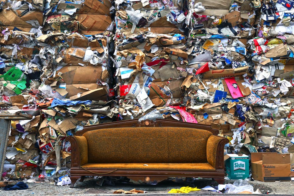 A vintage-style wooden sofa with ornate carvings and brown upholstery sits in front of a large wall covered with tightly packed, overflowing piles of crumpled cardboard boxes, paper waste, and packaging materials. The scene appears cluttered and dirty, with scattered debris and unorganized refuse overflowing onto the ground. The lighting is natural, highlighting the contrast between the detailed, worn sofa and the chaotic, trash-filled background. This image emphasizes waste accumulation and the importance of proper waste management and cleaning, as discussed on the West Hampstead furniture disposal and cleaning waste rules page at westhampsteadcarpetcleaning.co.uk, by professional cleaning services.